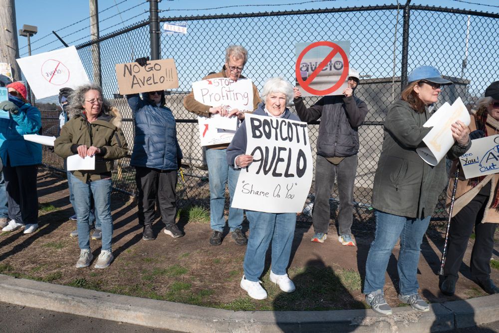 At the Avelo protest a group of people hold protest signs. The most obvious one in this picture reads, "Boycott Avelo." It is held by a grey haired woman.
