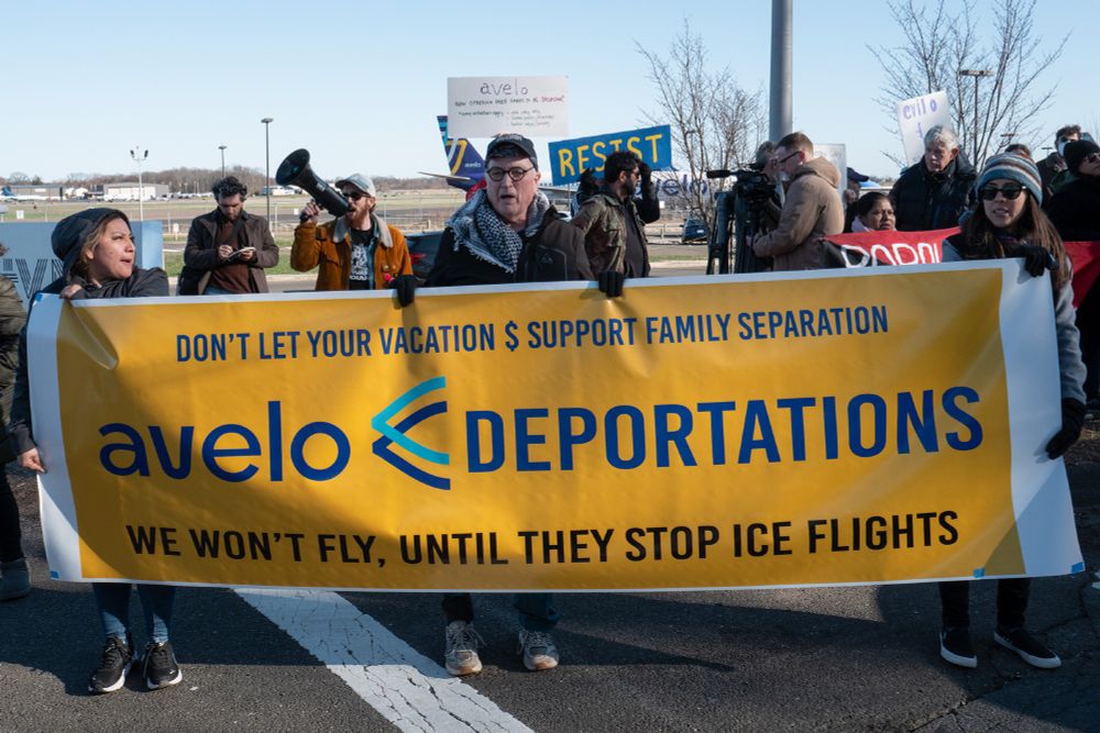 People in front of Tweed new Haven Airport with a large yellow sign big enough that three people span its length the hold it. In blue block letters against a yellow background it reads, Avelo Deportations. We won't fly until they stop ICE flights."