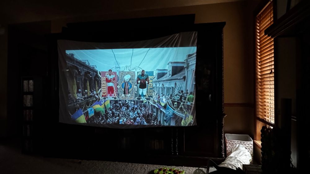 Picture of projection screen stretches across an entertainment center with a scene of a large crowd people on a street between two buildings and overlayed is a graphic with Patrick Mahomes over a light red background with CHEIFS spelled bottom to top in the lower left corner, a graphic of a Super Bowl trophy with a stylistic background, and a graphic of Jalen Hurts over a light green background with EAGLES spelled bottom to top in the lower right corner of the graphic box.