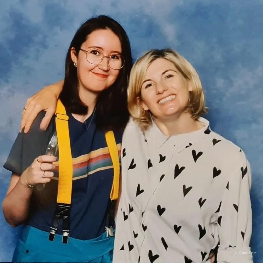 Cosplayer dressed as the thirteenth doctor stabbed with actor Jodie Whittaker. The cosplayer is wearing a dark blue top with rainbow stripe across the chest, tea trousers, and yellow suspenders. She has dark hair and is wearing round rimmed glasses and holding the thirteenth doctor's sonic screwdriver. Whittaker is wearing a white shirt with black hearts all over it. Whittaker has her arm thrown over the cosplayers shoulders. They are standing in front of a blue background.