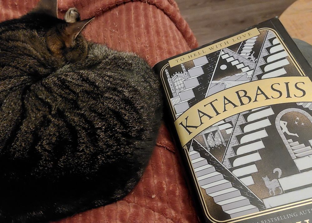 Een slapende kat op een rode deken met daarnaast het boek Katabasis van RF Kuang