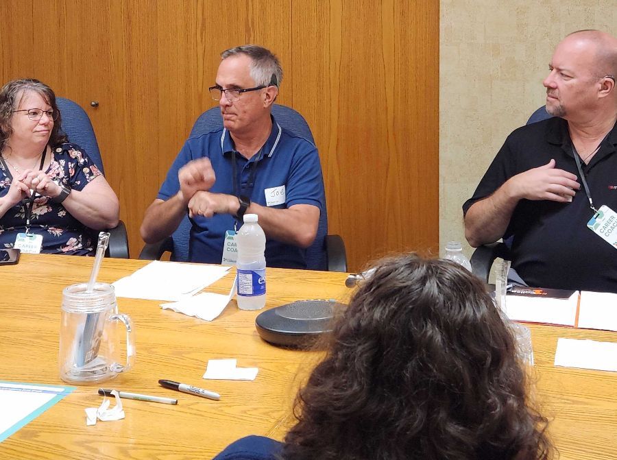 A second man gestures in ASL while others beside him watch with the back of a student's head across the table from him.
