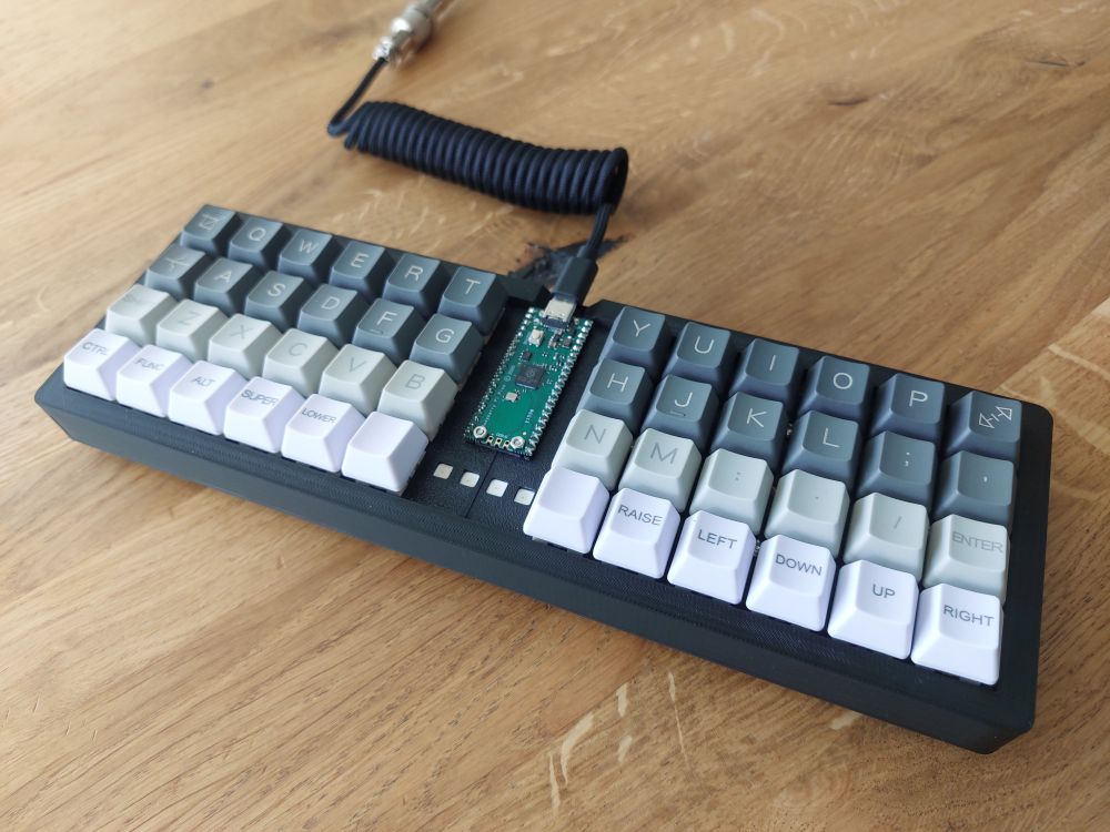 A view of the fully assembled keyboard on a wooden table. A coiled USB C cable plugs into an exposed raspberry pi pico board which is mounted to the top plate of the keyboard. On either side of the pico sits 24 keys - a distance of about 3cm apart. The keycap set is grayscale, with the bottom row of keys all being white, and the next 3 becoming progressively darker in value. Under the pico there are 4 WS2812 LEDs used for status.