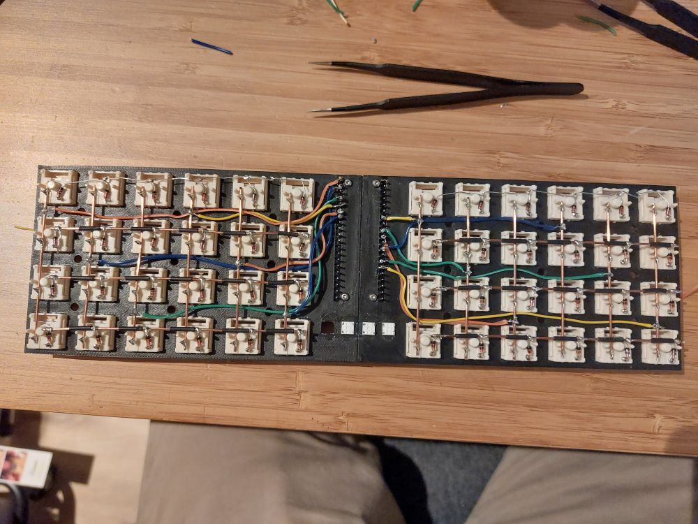 An in-progress view of the keyboard being built. The plate is shown upside down with all 48 keys pressed in. The keys are connected together in a hand-wired matrix, with the main backbone of the matrix being made of solid 1mm copper wire. The rows and columns are connected to exposed pins of the raspberry pi pico module with somewhat questionable soldering. The left side is done, while the right side is not yet complete. The RGB LEDs are not connected at all, and the leftmost one is missing entirely. 