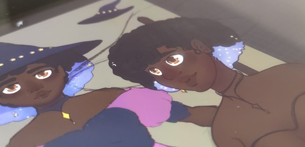Photograph of a character design in progress the character is a person of color with orange brown eyes, a black Afro with blue under neath and accessories of stars and a moon clipped in. They appear to wear a blue and pink top and a blue witch hat.