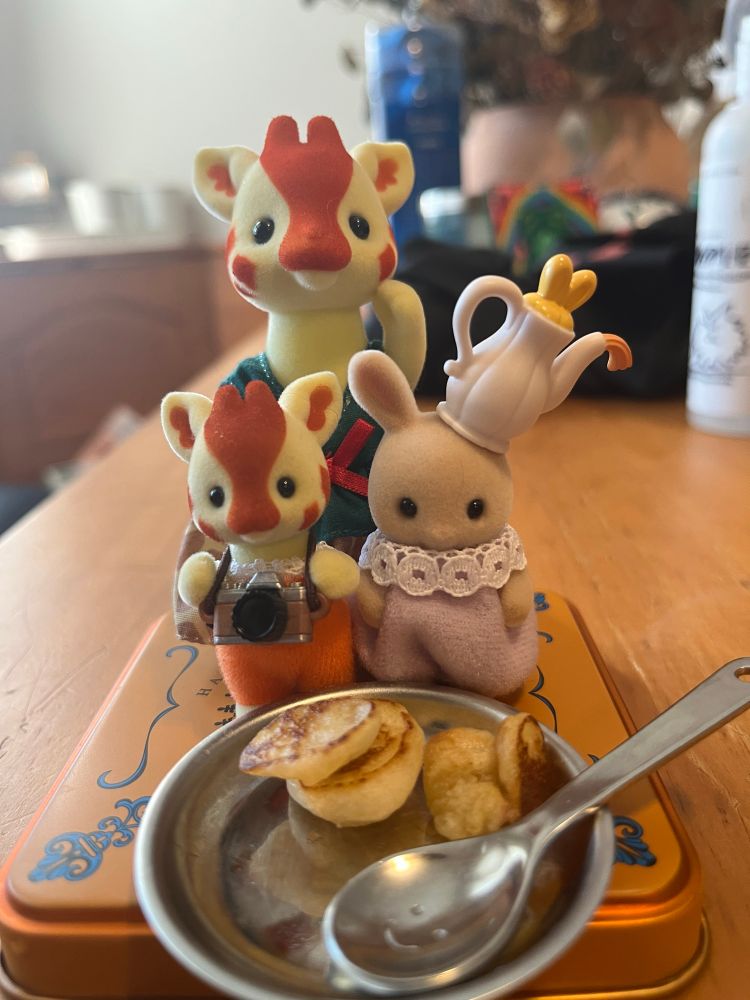 Three toys shaped like an anthropomorphic giraffe mama, a giraffe baby with a camera, and a baby bunny with a teapot on its head for a hat enjoy some pancakes for breakfast. 