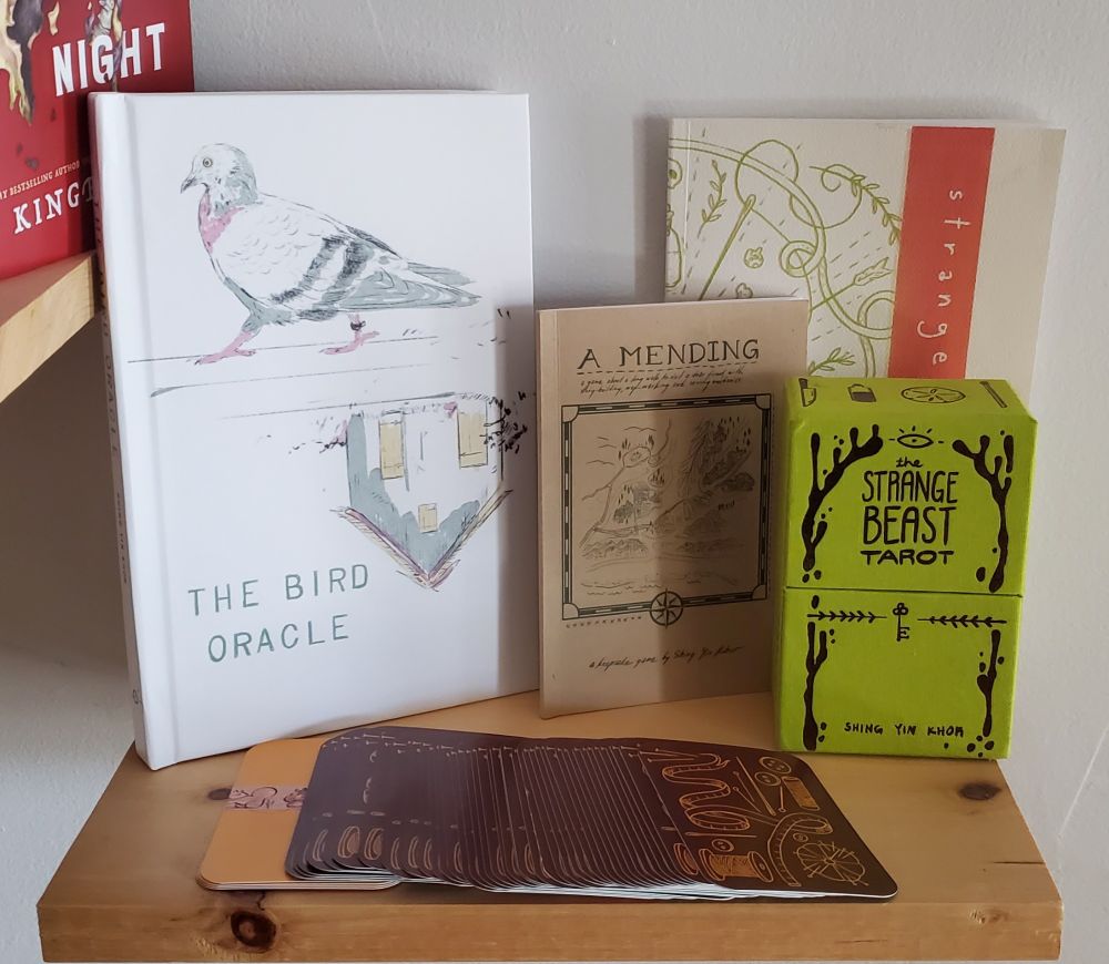 A photo of several art projects by Shing Yin Khor: their latest game book The Bird Oracle which features a pigeon walking and a house upside down under it both in cool gray, pink and yellow on a white background. Next to it are the booklet and cards from A Mending (a game about a long walk to visit a dear friend). Next 
to them is the Strange Beast Tarot deck in a beautiful green fabric box with gold accents, the booklet for the tarot deck is visible behind the box. 