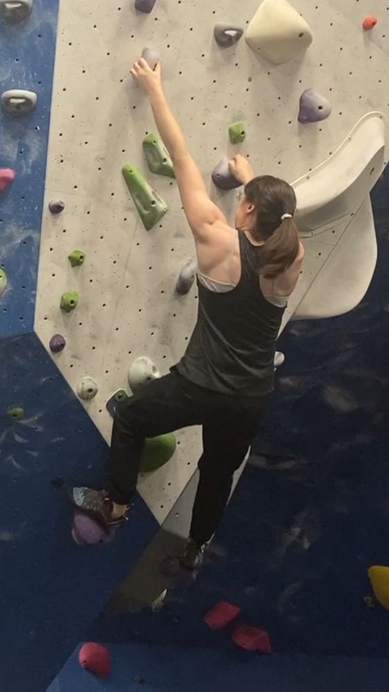 She’s using momentum from the last move to reach up to a higher hold with her other hand. She’s wearing a racerback tank top that shows off her shoulders