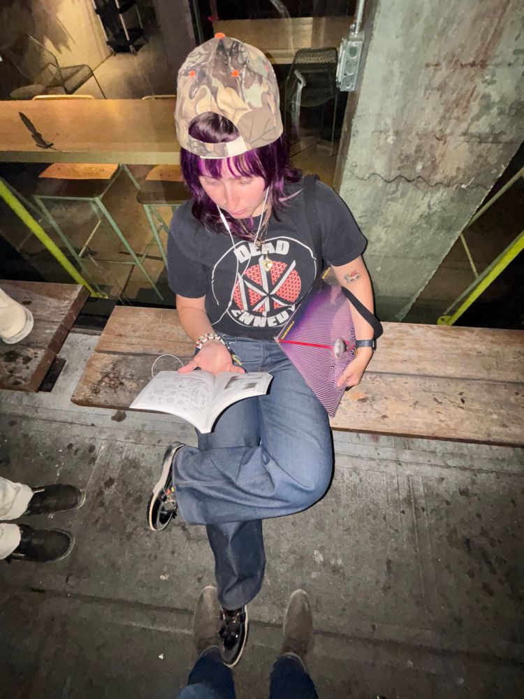 A photo of a nonbinary individual wearing a camouflage hat, a Dead Kennedys tee, raw selvedge denim pants, and loafers while looking at a page from the manga “Nana” and holding a vinyl of “Currents” by Tame Impala. They are also wearing headphones that aren’t plugged in to anything.