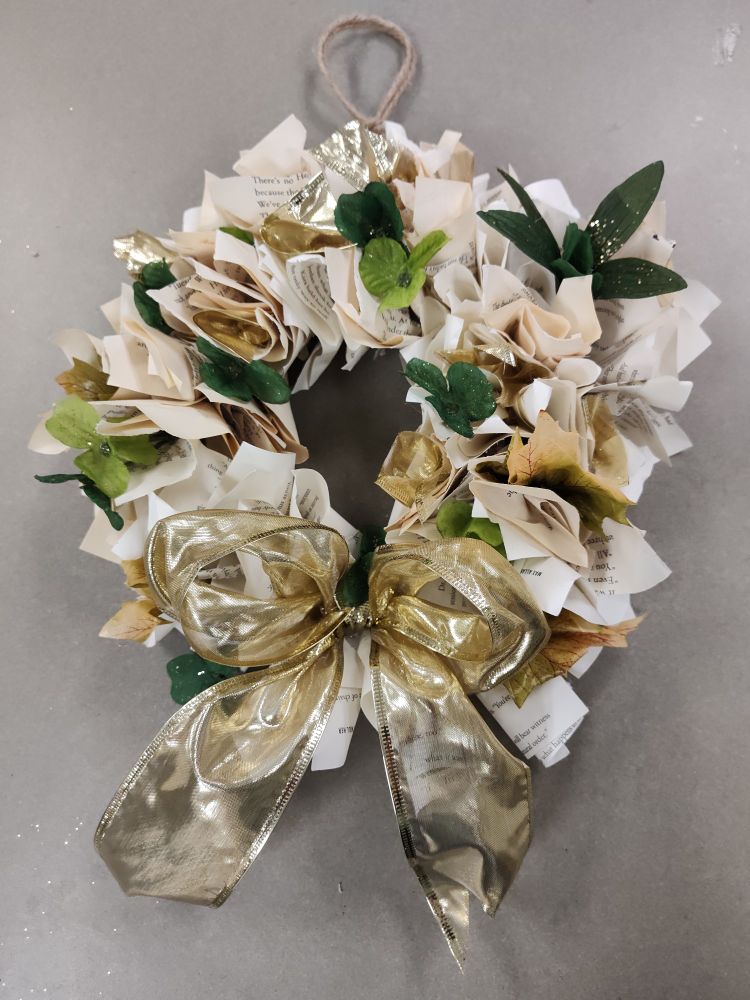 A homemade wreath using pages of books. There is some green foliage and a golden bow for pops of color.