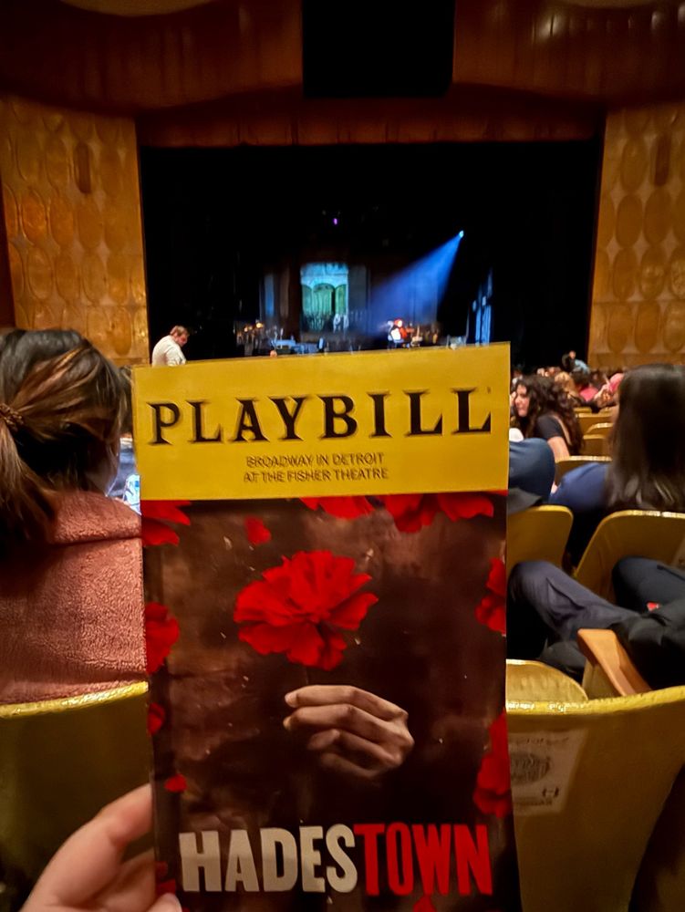 A Playbill for "Hadestown" held up at the Fisher Theatre in Detroit, showing its distinctive dark cover design with red carnations. The historic theater's ornate gold interior and stage are visible in the background.​​​​​​​​​​​​​​​​