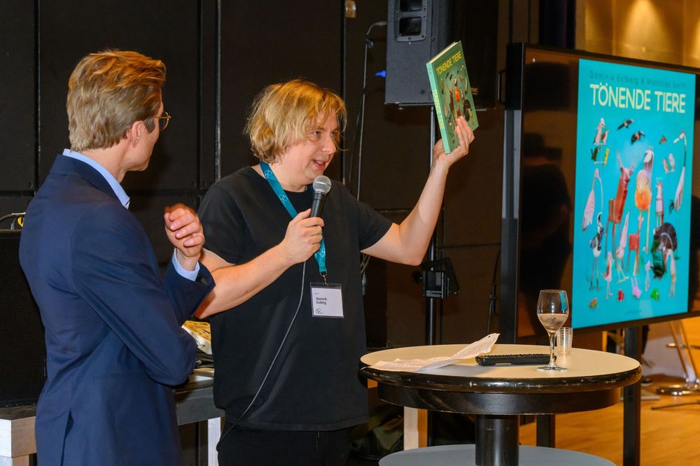 Zwei Personen auf Bühne vor Display, der Vögel zeigt. eine Person hält ein Buch hoch