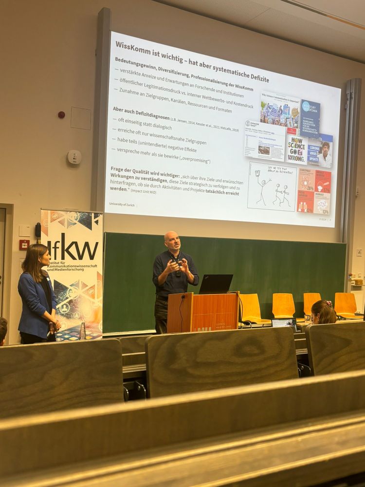 Zwei Personen präsentieren in Hörsaal Folie über Bedeutung und systematische Defizite von Wisskomm