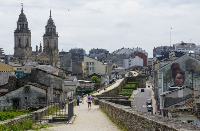Part of the pathway at the top of the Roman wall which encircles the Galician city of Lugo in Galicia. it is at quite an elevated position above a road to its right. The path is roughly sand in colour and is quite sinuous but appears to take a sharper curve to the left in the distance. To the left is the inner city with two church steeples being the most prominent features. The side wall of a house is visible to the right of the Roman wall with an indistinct mural of a young girl.