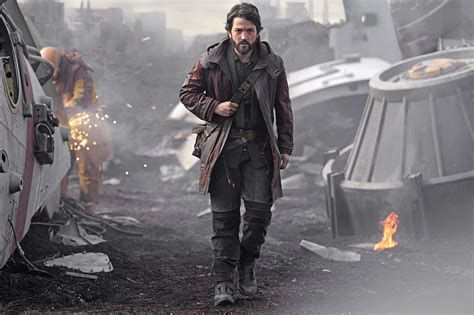 Cassian Andor, lead character of Star Wars "ANDOR" (played by actor Diego Luna), walking toward camera in dark clothes, looking determined. He wears dark clothes, including dark shirt, pants, boots, and a reddish-brown, possibly leather, mid-thigh-length jacket. He carries a bag or satchel that is barely visible behind his right hip; its strap crosses his body from left shoulder to right hip, held by his right hand.