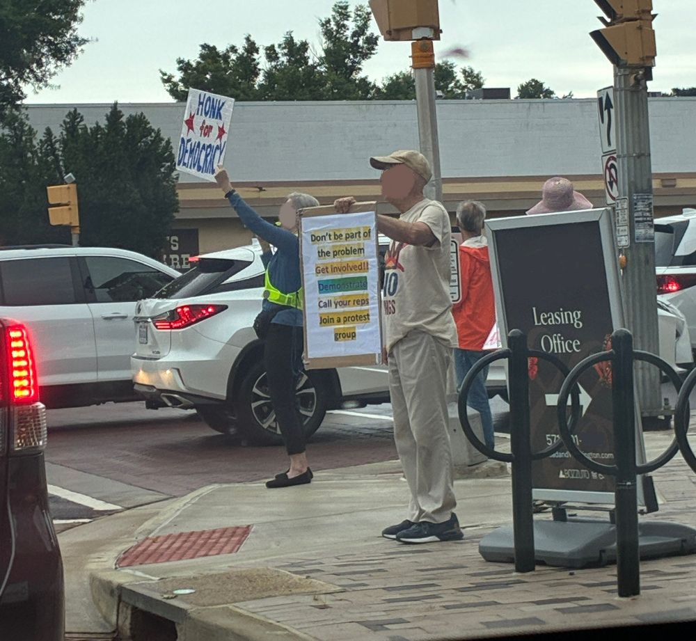 3-4 Protesters standing with signs on a street corner. Beyond them are 2 lanes of cars stopped in traffic. Woman wearing a neon safety vest holds up a sign reading "Honk for Democracy". Man in a "No Kings" tshirt carries a sign reading "Don't be part of the problem. Get involved!! Demonstrate / Call your reps / Join a protest group. Other protesters and signs are obscured from view.  Faces are blurred, because I don't know them and didn't ask permission to post photo.  
