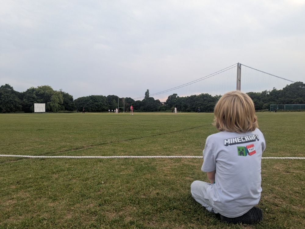 It's Hessle cricket club, under the shadow of the Humber Bridge. A young child with long hair is sat in the foreground, watching a game