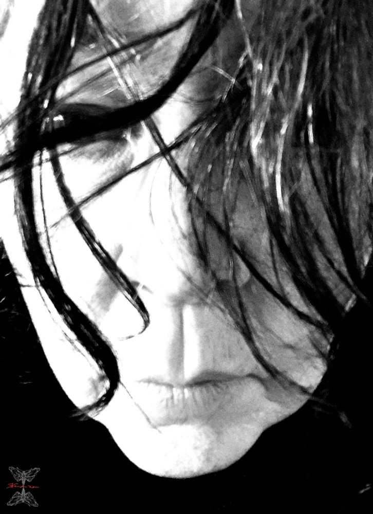 Photo en noir et blanc. Visage flippant de l'artiste qui a le covid. Visage blafard, regard caché derrière des mèches noires et blanches. Fond noir. Un seul œil est visible. 