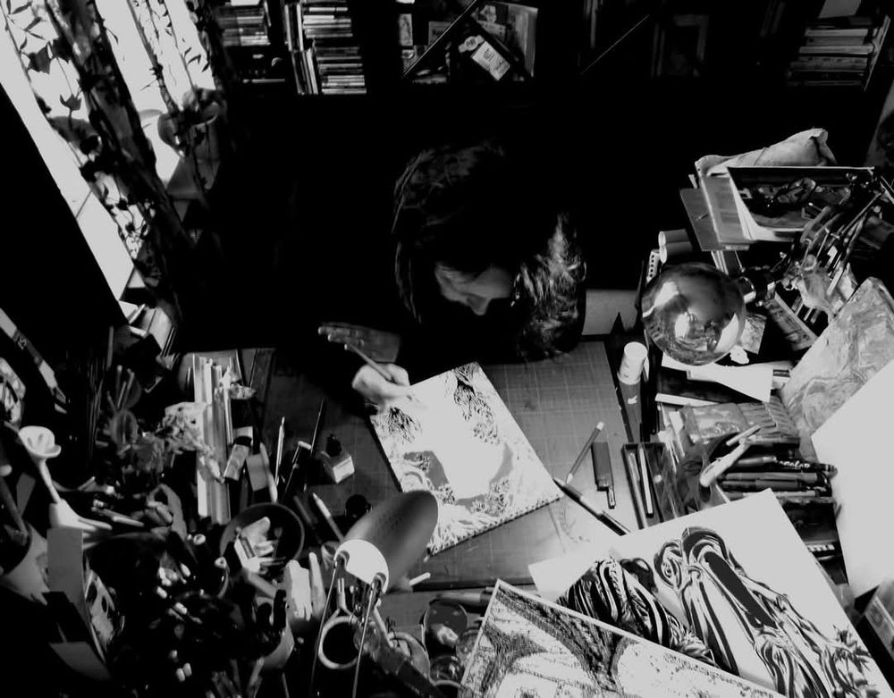 Artiste au travail. Table très encombrée, matériel, dessins, livres.