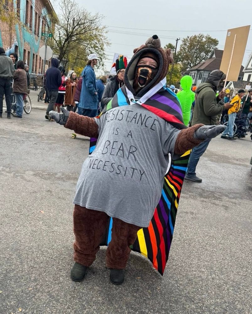 A Bear with a rainbow cape at the No Kings rally in Missoula, Montana with a shirt saying, “Resistance is a Bear Necessity”