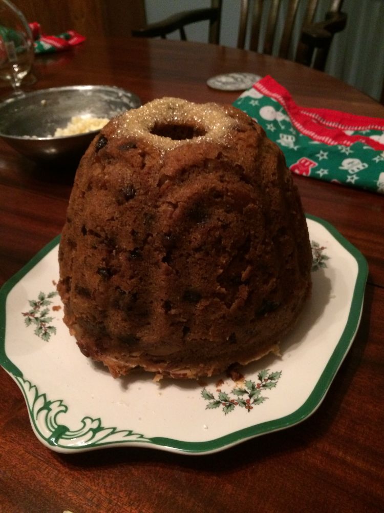 Tall Bundt -ish shaped cake, with textured arched lines from the pan. Golden brown in color with speckles of fruit throughout. Ring of brown sugar around the hole in the top (Which will caramelize later when it’s doused in alcohol and set on fire). Sitting on a square Spode Christmas cake platter with holly leaf and berries in the pattern and a green stripe along the edge