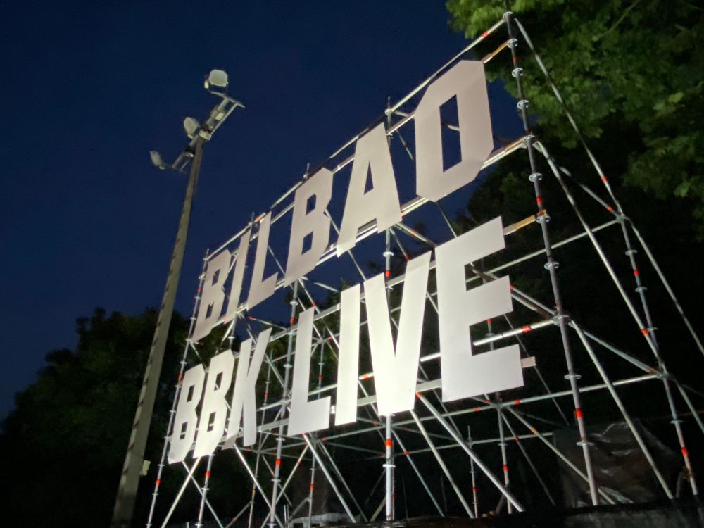 Photo taken at night, showing the name of the festival (Bilbao BBK Live) in huge letters, Co strutted on a large scaffolding structure. The structure is light up with bright lights.