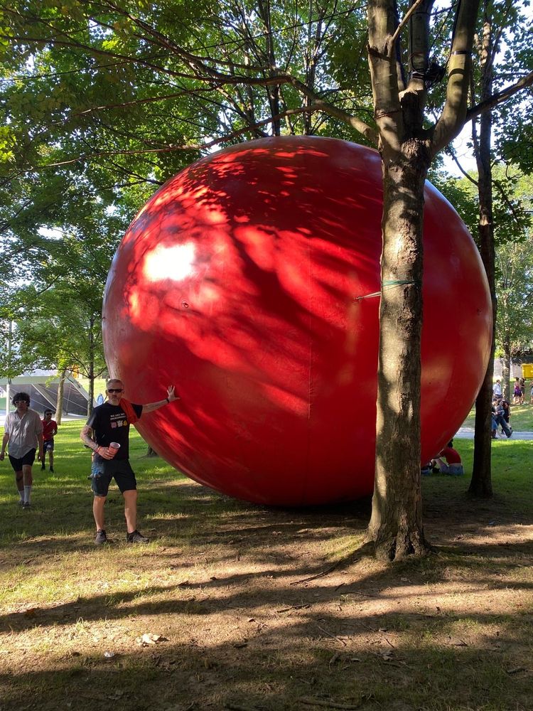 This is a photograph of the Bluesky account holder, Blakey_Bloke, standing next to the RedBall, a bright red 4.5 m diameter inflatable vinyl sphere. It is an art project of the American artist, Kurt Perschke. The RedBall appears in locations all over the world, usually ‘jammed in’ somewhere, and tethered into place. It commonly appears for a day, a couple of days at most. The list of locations that it will make future appearances at is not publicly disclosed. The project has been running since the early 2000s.