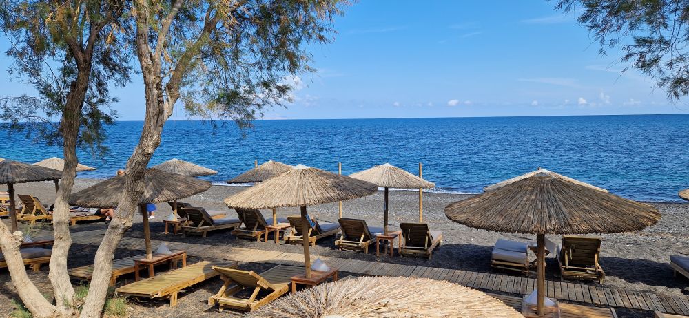 Beach of Kamari, Santorini, Greece. A black sand beach with lots of wooden sunlounes and sun shades made of straw. Large olive trees also provide shade. The beach is almost empty. Very beautiful and idyllic scene.