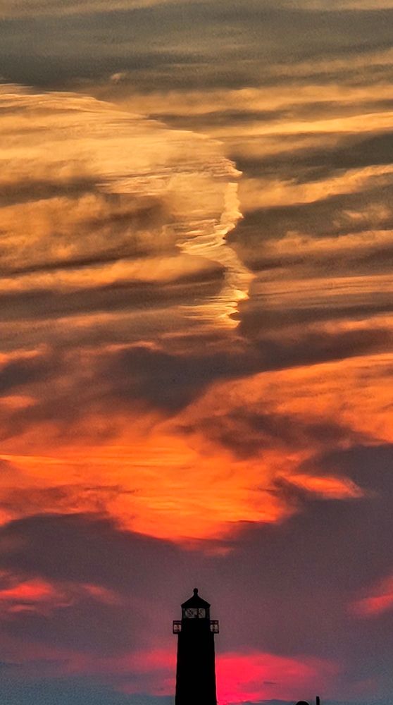 The dark silhouette of a tiny lighthouse sits centered at the base of a vibrant sunset’s portrait as fiery layers of night sky get extinguished by the horizon.