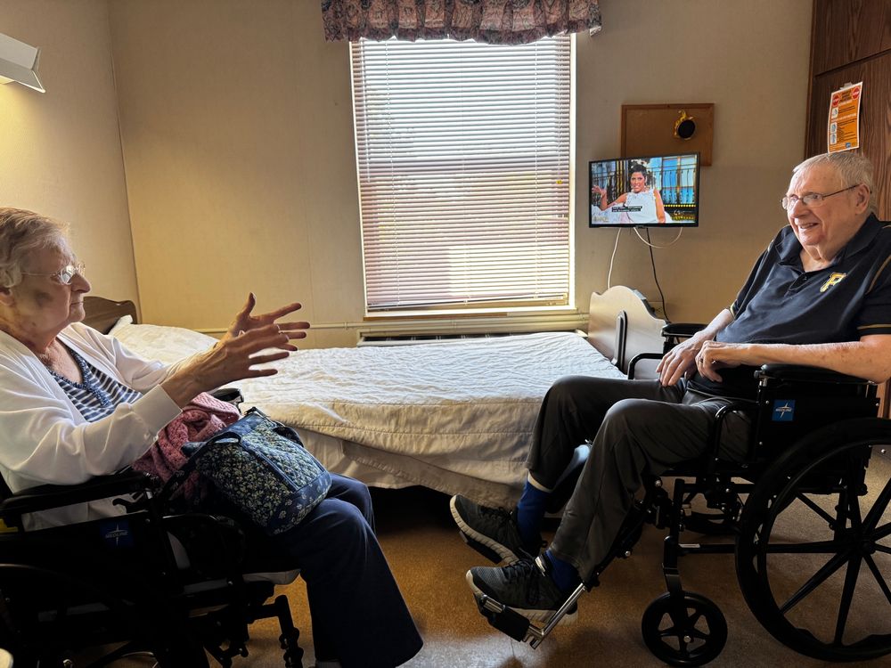 Mom and dad sitting in wheelchairs facing each other talking and smiling. Rehab facility bed in the background 