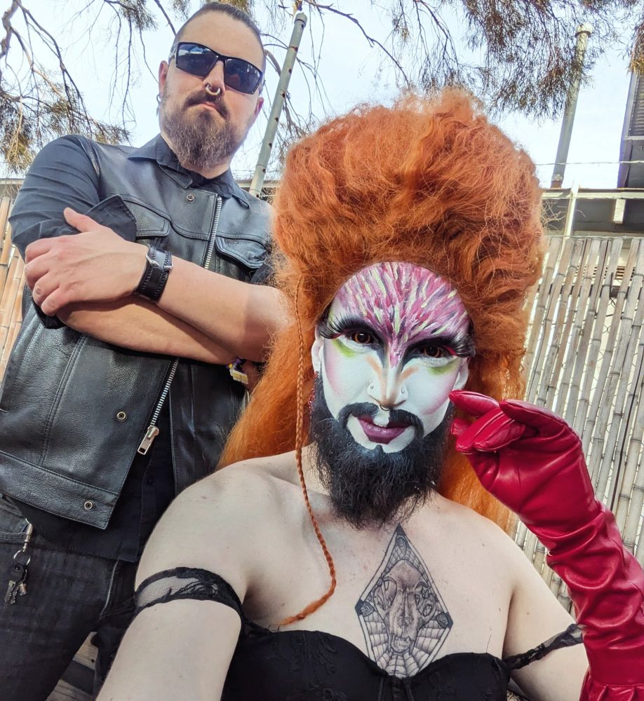 A drag queen with a clown white face, long beard, blacked-out eyes, and large orange hair sits in front of a standing, bearded man in sunglasses and leather attire. Both are smirking at the camera.