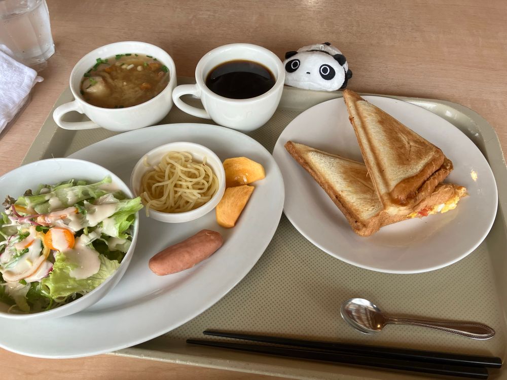 cafeラ・セルのホットサンドモーニングセット。
卵とトマトのホットサンド、ボウルに入ったサラダ、ペペロンチーノ風パスタ、ソーセージ、柿、お味噌汁、コーヒーがプレートに乗っている。
それを見つめるたれぱんだのぬいぐるみ。