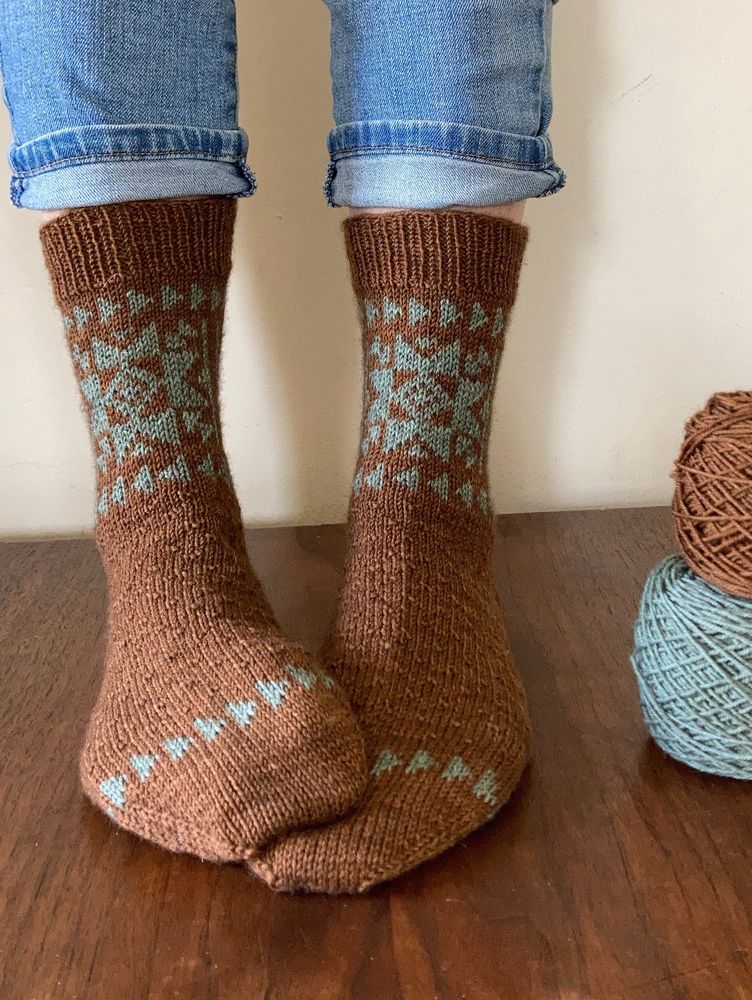A closeup of feet wearing copper brown calf-length socks with sage green sawtooth stars on the leg and flying geese triangles on the leg and above the toe. The top of the sock foot has a quilt-inspired texture.