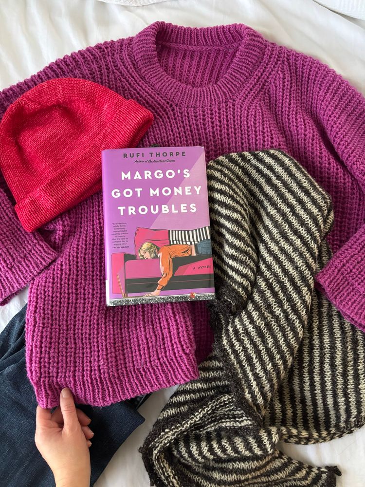 A book surrounded by handknits that match the cover: hot fuchsia sweater, bright pink hat, black and white striped shawl.
