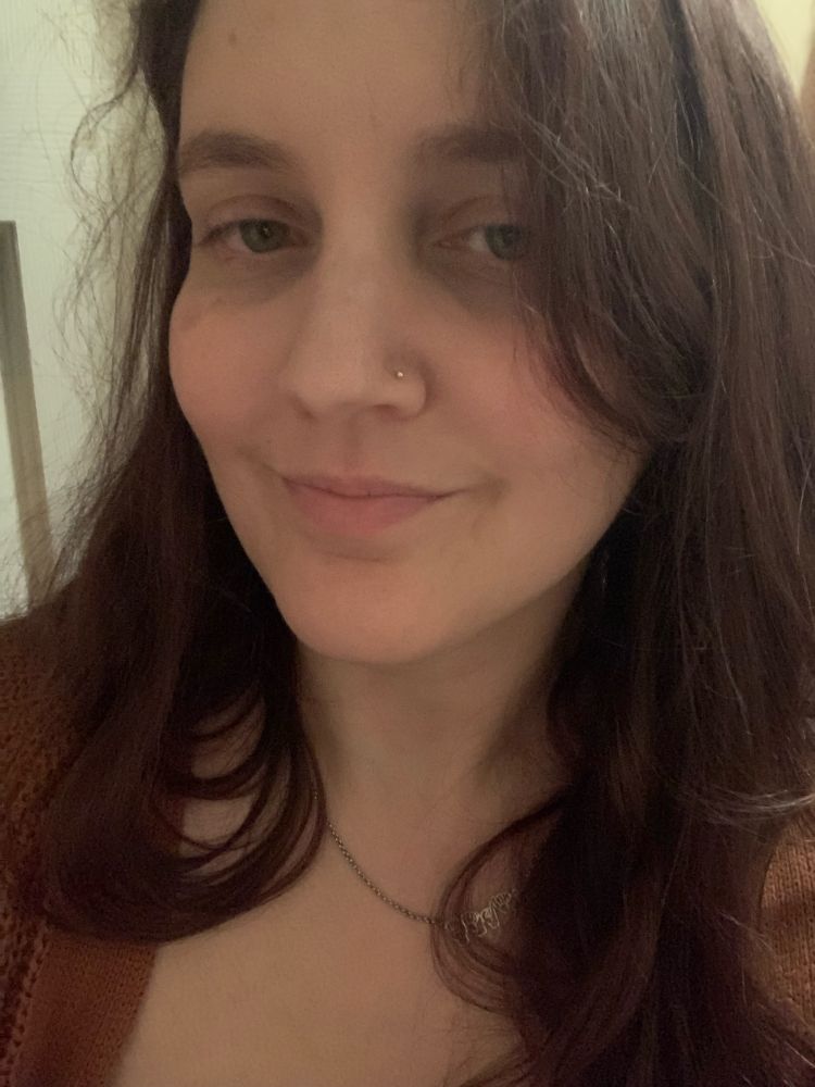 Tired femme looking white person with long brown hair wearing a light brown sweater 