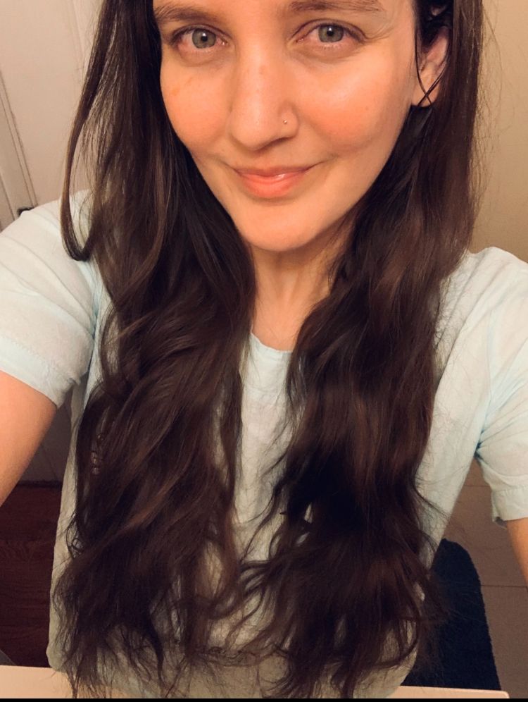 Femme person staring directly at  the camera. They have long dark brown hair and are wearing a light blue shirt 