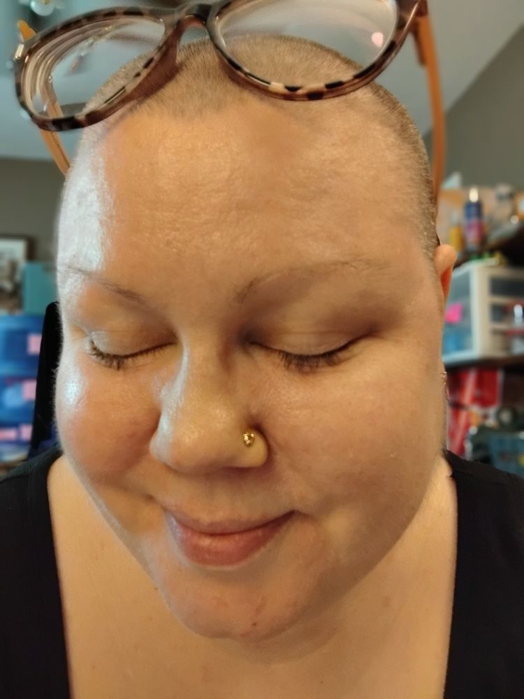Female person has eyeglasses resting on top of her head. Eyes are closed, and there is a small, gold ladybug stud nose piercing displayed.