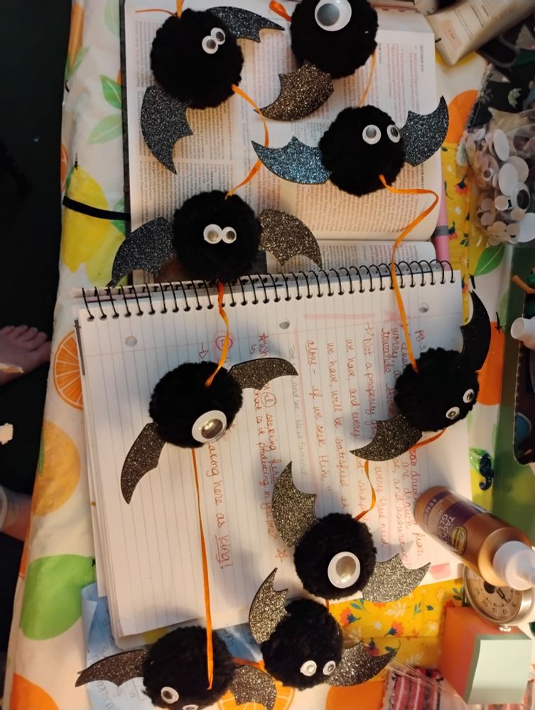 Black pom pom bats with sparkly wings, on an orange ribbon garland. Googly eyes of varying sizes are affixed in varying arrangements. Even a cyclops bat or two. Spooky season approaches.