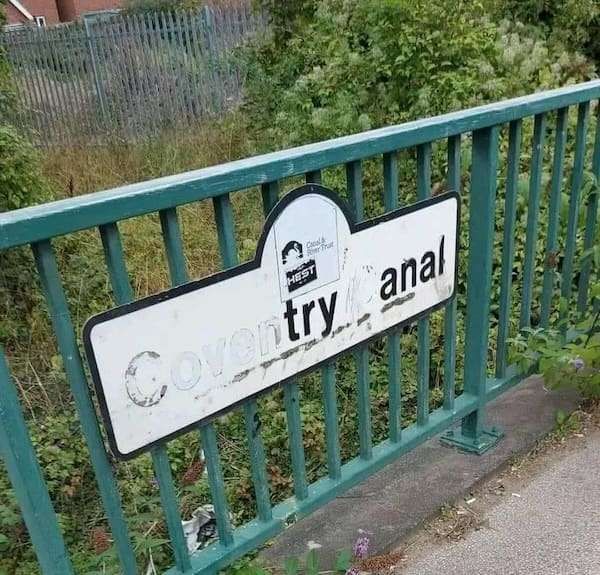 black and white sign for 'Coventry Canal' on a green fence. most of the letters are faded so it appears the sign says "try anal" instead.