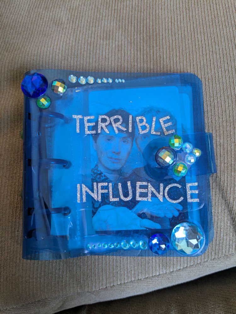 A small photo album/binder that is blue. There are gems in the top left and bottom right corners and gems along the top and bottom of the cover. There is a little button to hold it closed and there are gems there too. There are also sparkly letter stickers in the middle that day "Terrible Influence"