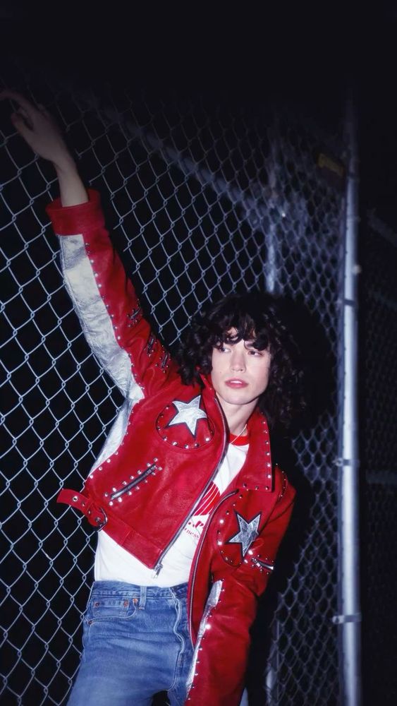  Conan Gray leaning against a fence with his left hand reaching up and holding onto part of the fence. He is wearing a red leather jacket and a white t-shirt underneath and blue jeans.