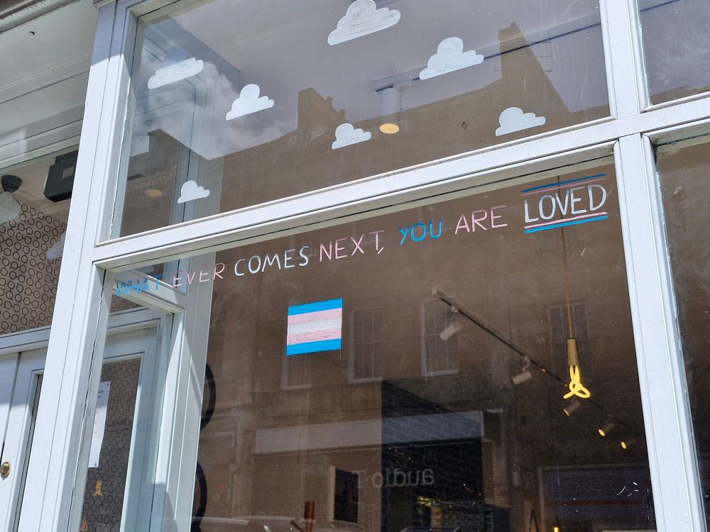 'Whatever comes next, you are loved' written in a window in the trans flag colours. 