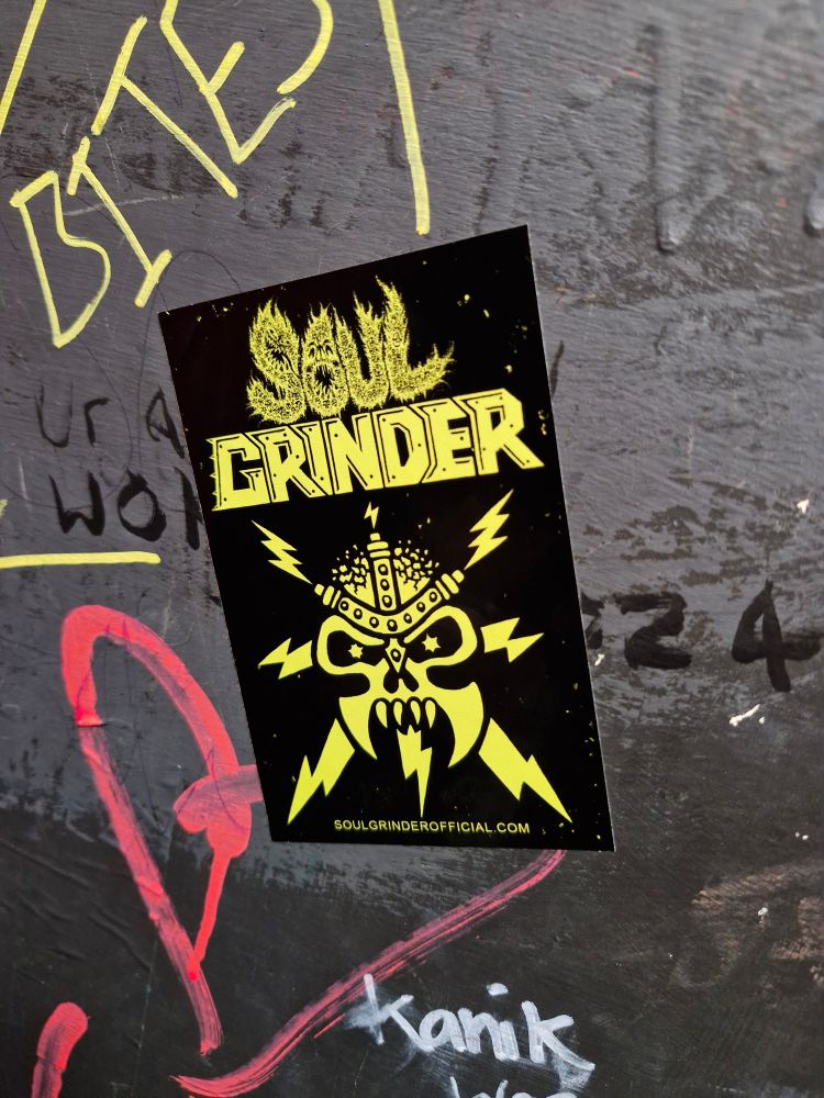 A sticker from the band Soul Grinder stuck to a wall which has graffiti underneath. 