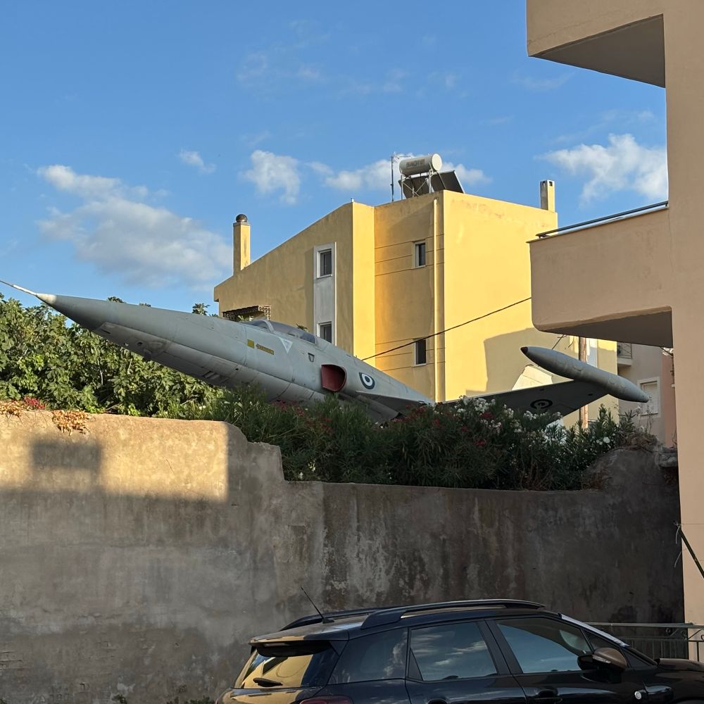 An old fighter jet is visible above a garden wall in a residential area of Kalamata, Greece.