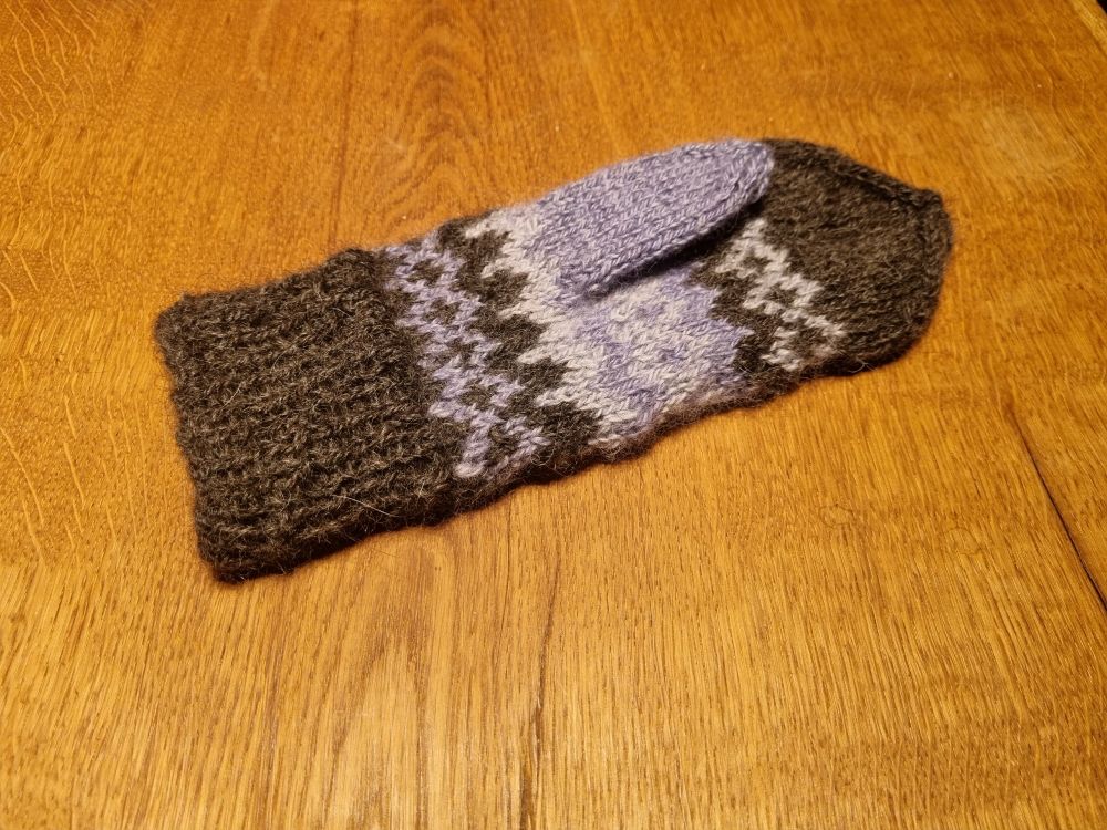 Ein fertig gestrickter Fäustling mit grauem Bund, grauer Spitze und dazwischen Muster in Lilatönen und lila Daumen liegt auf einem Holztisch