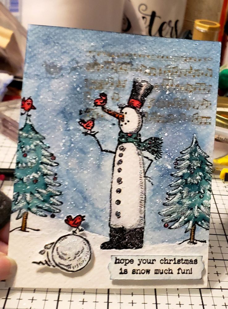 Christmas card with a snowy scene of a tall snowman holding a cardinal bird with another cardinal perched on his carrot nose singing.  Evergreen trees on either side of him. One has a cardinal standing on the tip top of the tree.  Trees are adorned with red Christmas balls.  Another cardinal is rolling by atop a snowball.  Snow is falling. In the background  Santa on his sleigh being led by his reindeer is in faint silhouette. Text says, "hope your Christmas is snow much fun". They all appear to be having a good time! 