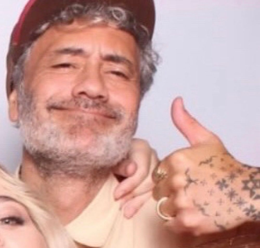 Taika giving a thumbs up with his left hand. He’s writing a beige shirt and a brown hat. 
