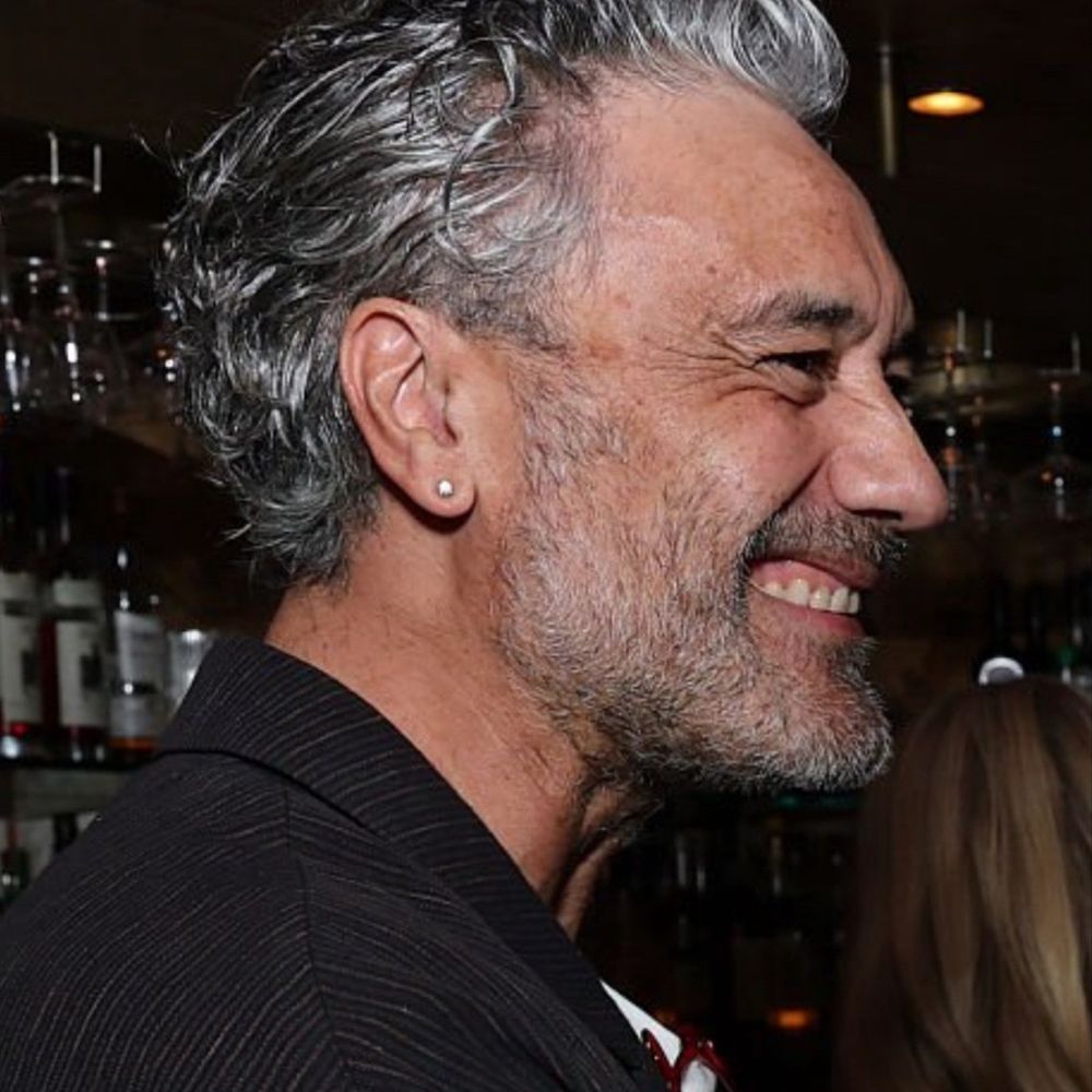 Taika at a club. We’re seeing him from the side and he’s got a big smile. He’s wearing a black blazer with tiny dark grey vertical stripes. 