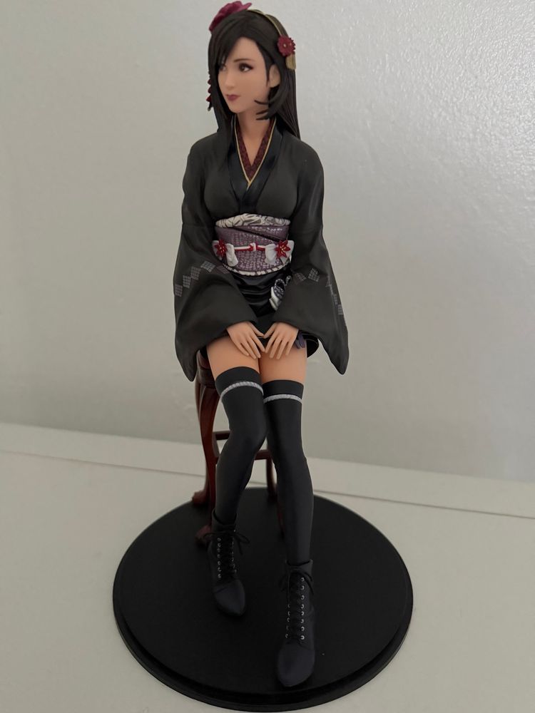 Anime-style figurine of Tifa from Final Fantasy VII Remake sitting on a chair. She has straight, dark hair adorned with red hair ornaments and is wearing a black kimono with a patterned sash (obi) featuring floral and geometric designs. The kimono sleeves are loose, and she has her hands gently folded on her lap. She wears thigh-high black stockings with white bands near the top and black lace-up boots, and sits on a circular black base. The overall pose and expression are calm and elegant.