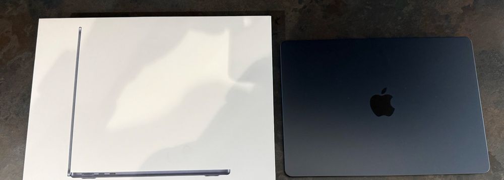 A rectangular white box and a closed laptop resting on a dark, stone-like surface. The box is at the top of the image. Its design is minimalistic, featuring a side-view image of a very thin laptop printed in the center. This image shows the left edge of the laptop with visible ports including a charging port and other connection slots. The box’s top left corner is slightly crumpled, showing a small crease.

Below the box is the actual laptop, positioned parallel to it and taking up the lower half of the image. The laptop is a very dark gray, almost black in color. It is closed and its surface is matte, with the iconic glossy black Apple logo centered on the lid. The corners of the laptop are rounded, and its profile is very slim, reinforcing its modern design. The lighting in the image highlights the smoothness and sleekness of the laptop's surface.